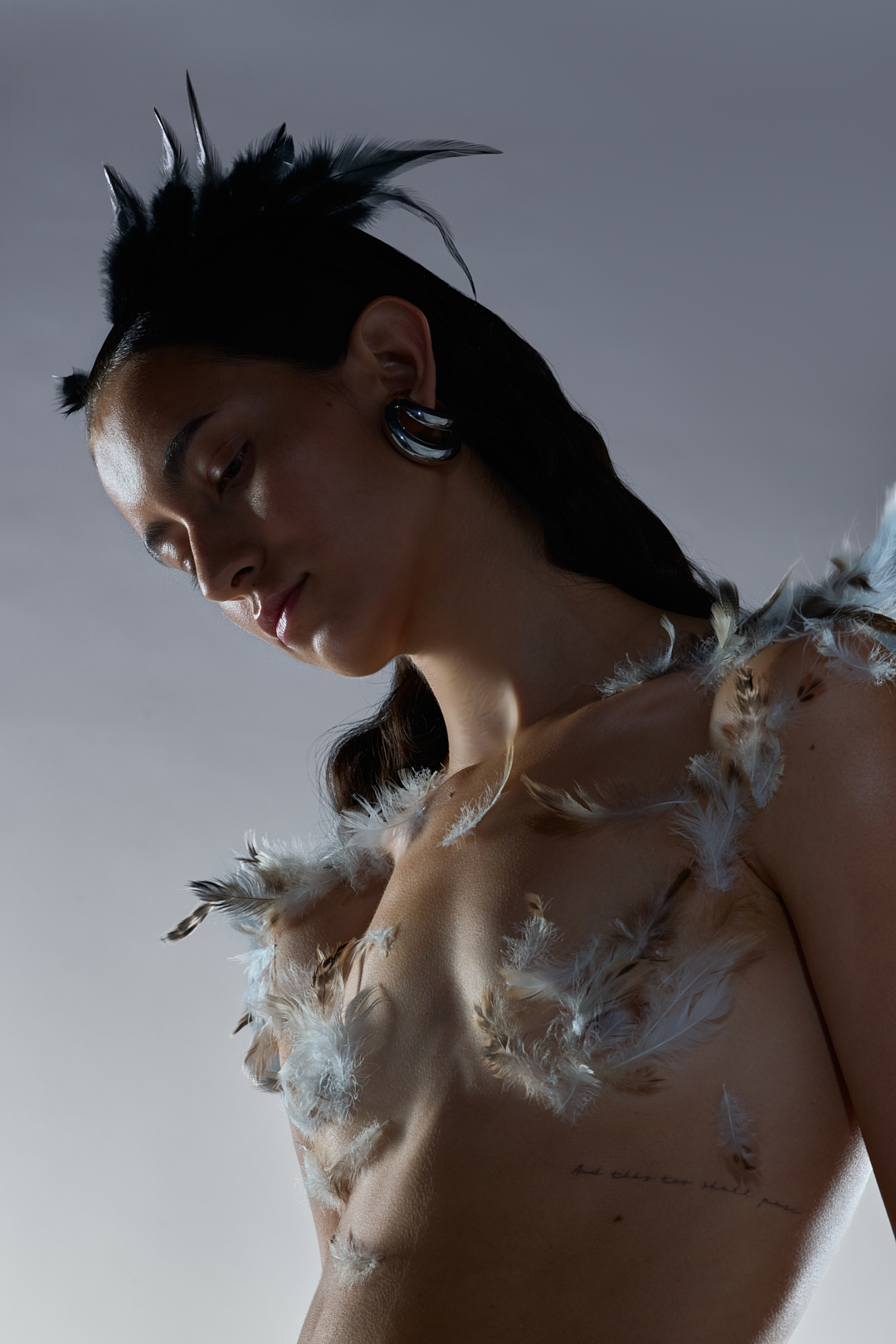 Close-up portrait of a woman adorned with feathers on her shoulders and a dark feathered headpiece against a neutral grey studio background