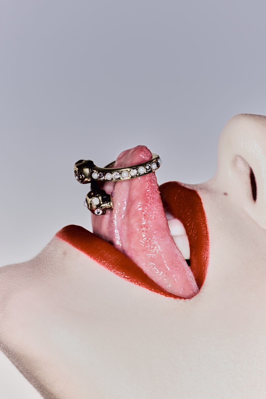 Close-up of a woman's face with a ring piercing her tongue. She has red lipstick on, and the background is a neutral color.
