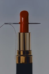 Red lipstick in a gold and black case pierced horizontally by a silver sewing needle and thread against a grey studio backdrop.