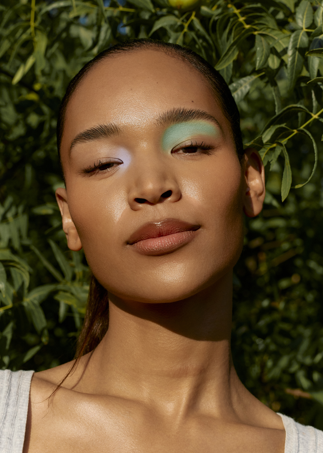 Close-up of a woman with artistic blue and lavender eyeshadow, posing outdoors surrounded by green leaves in soft, natural light.