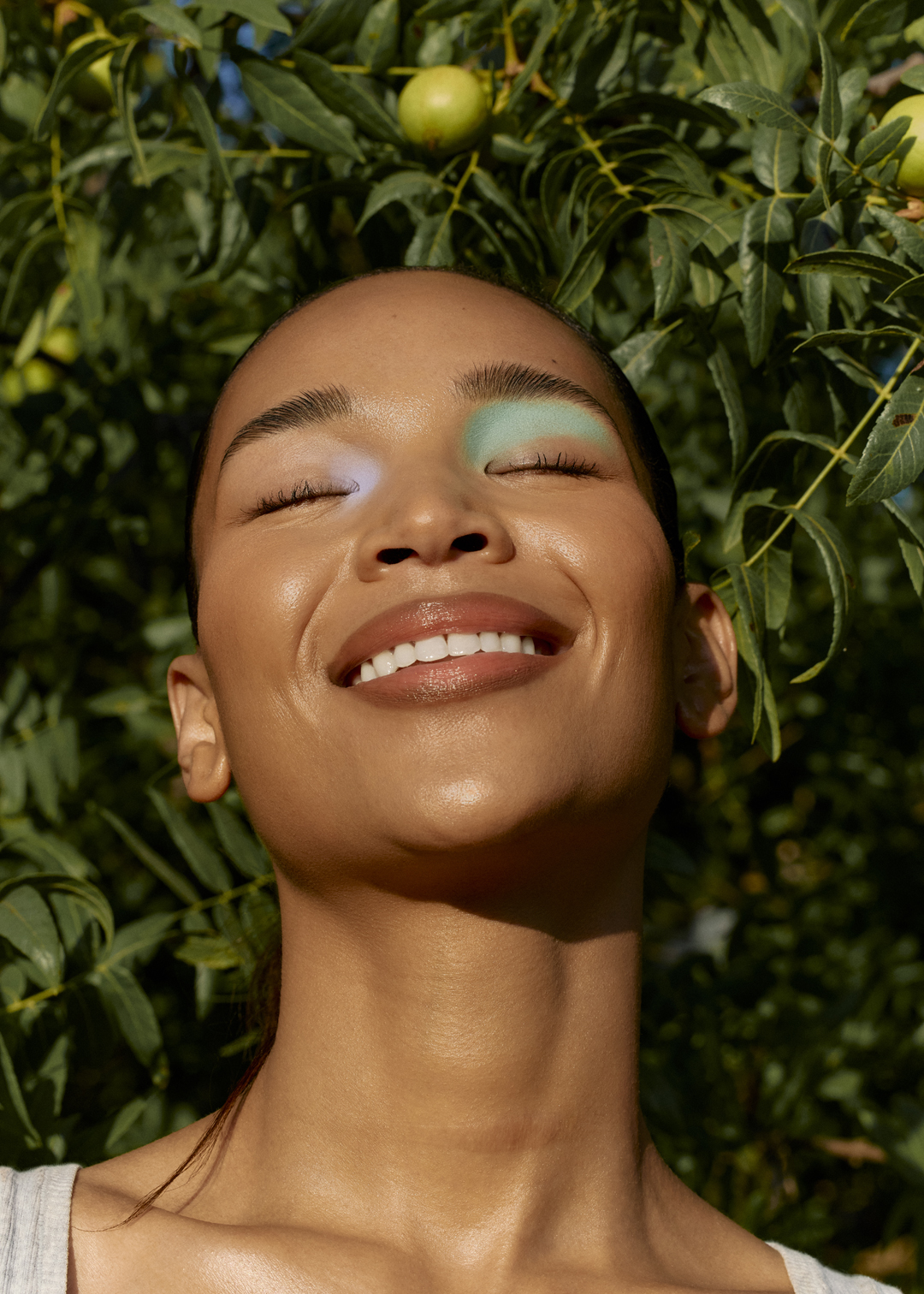 Portrait of a woman with blue eyeshadow, eyes closed, smiling outdoors surrounded by lush green foliage in a natural, sunlit setting.