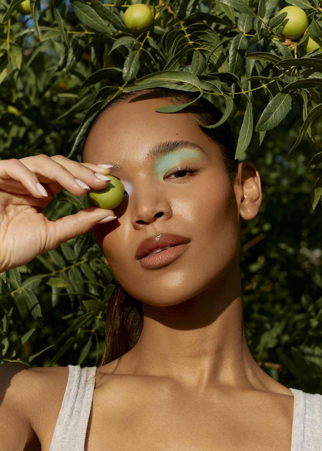 Portrait of a tanned woman with creative makeup holding a green fruit against a leafy green tree background in outdoor daylight.