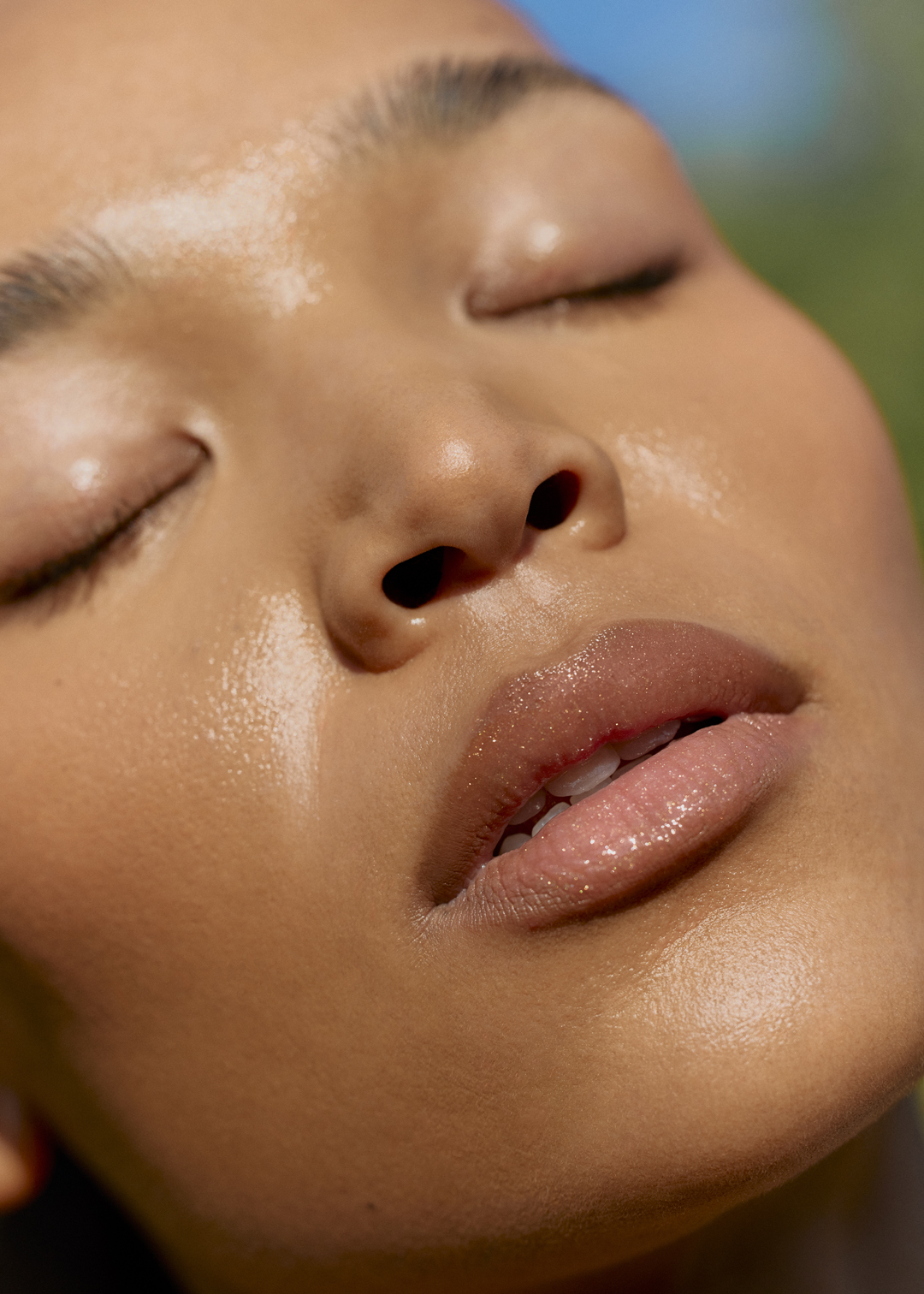 Close-up portrait of a young adult woman with tanned skin and closed eyes, outdoors in natural sunlight with a blurred green and blue background.