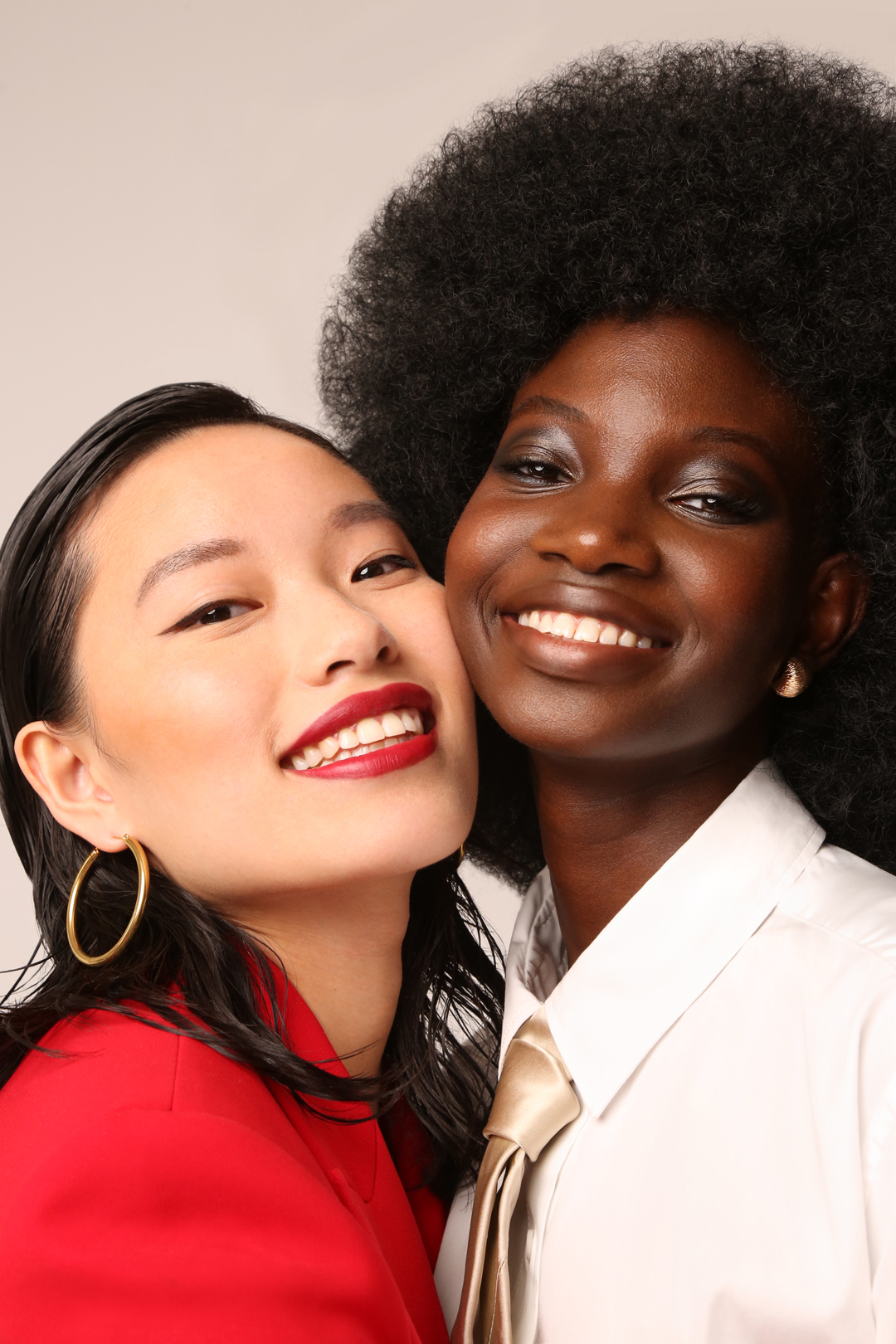 Close-up of two smiling women, one Asian with a red blazer and one Black with an afro, wearing makeup and formal wear, indoors.