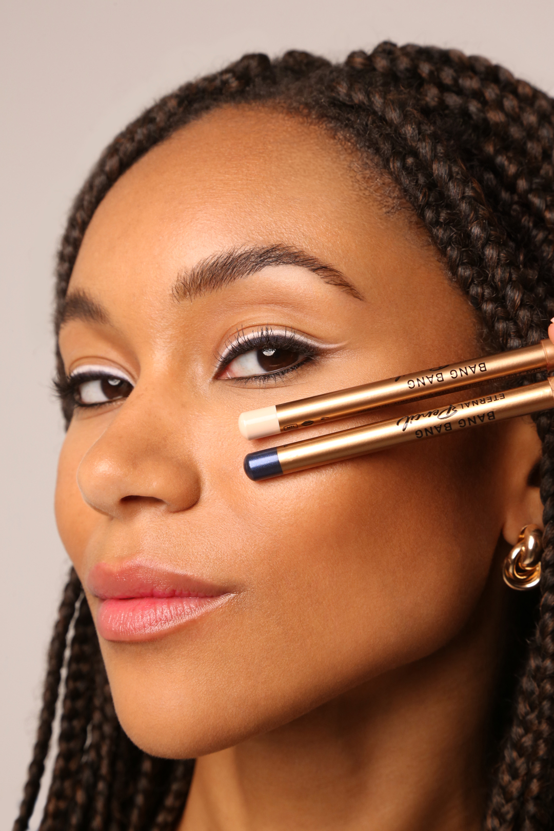 Close-up of a woman with dark braided hair modeling eye makeup using cosmetic eyeliner pencils in front of a neutral background.
