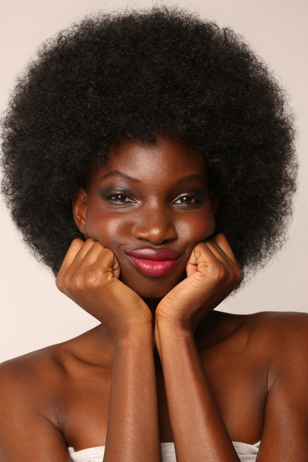 Close-up of a dark-skinned woman with an afro hairstyle, wearing makeup and a white wrap, holding her face with her hands indoors.