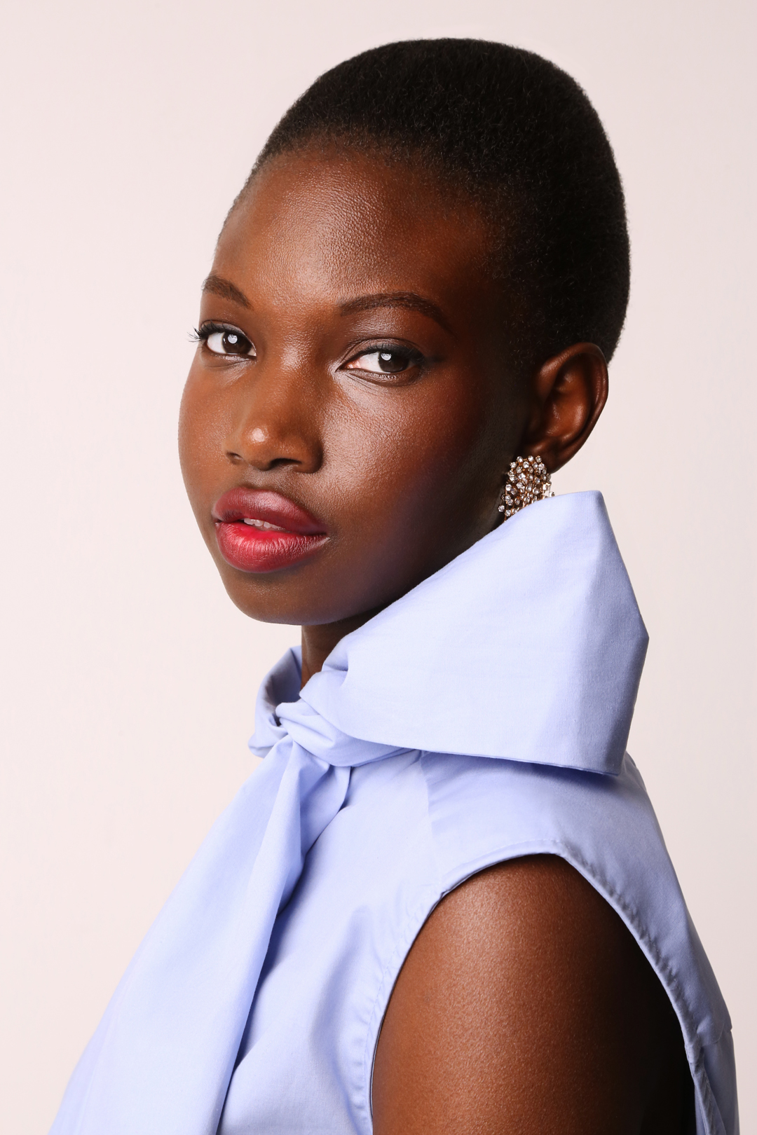 Close-up portrait of an African-descent adult woman with short hair, wearing a light blue top and earrings, against a neutral background.
