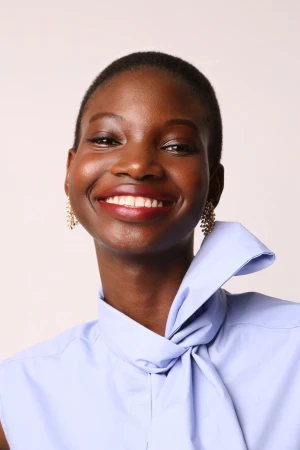 Portrait of a smiling Black woman wearing a light blue tied-collar top and earrings against a plain background