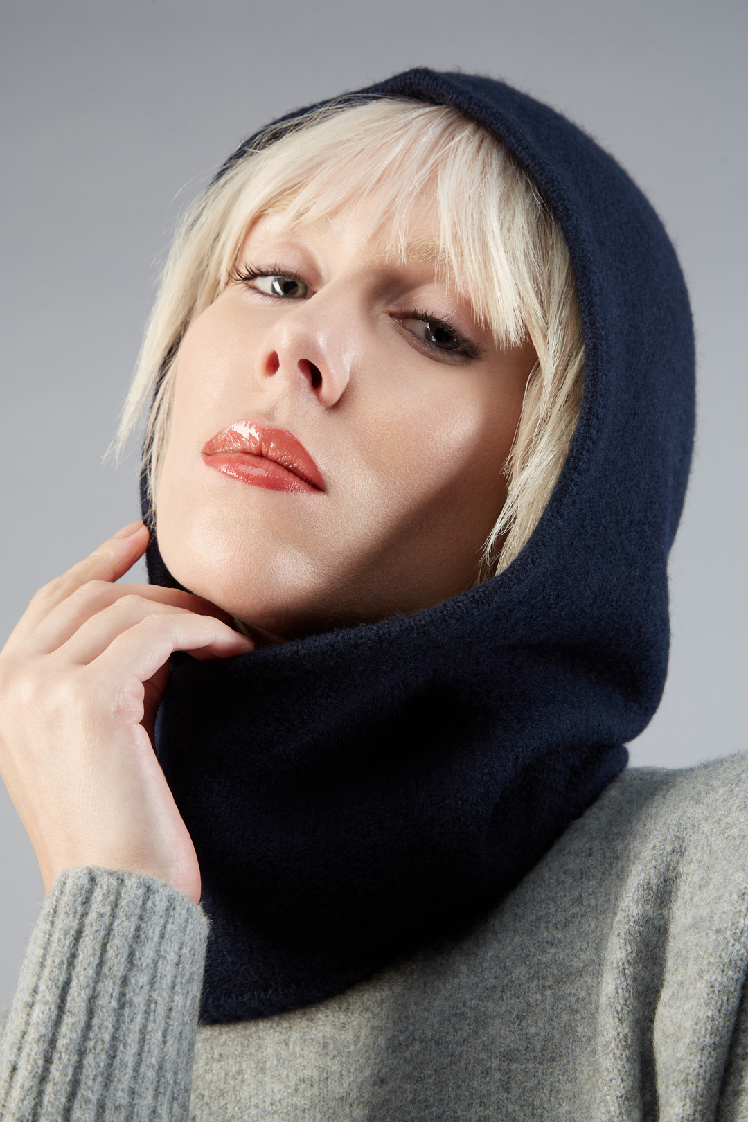 Portrait of a fair-skinned adult woman wearing a navy blue hooded scarf and a gray sweater, set against a plain gray background.