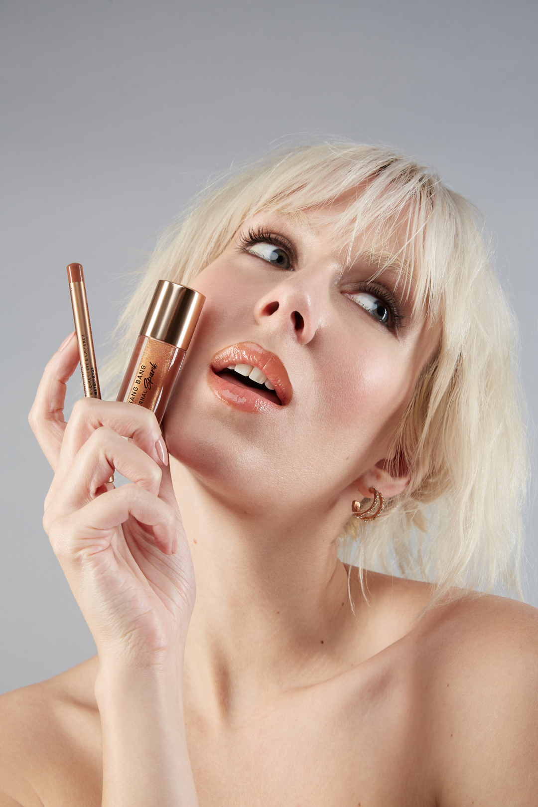 Close-up of a blonde adult woman holding makeup including lip gloss and lip liner against a gray background in a beauty shot.