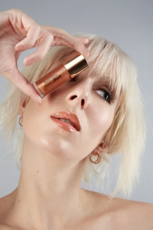 Close-up portrait of a blonde woman holding a cosmetic product tube near her eye against a grey studio background