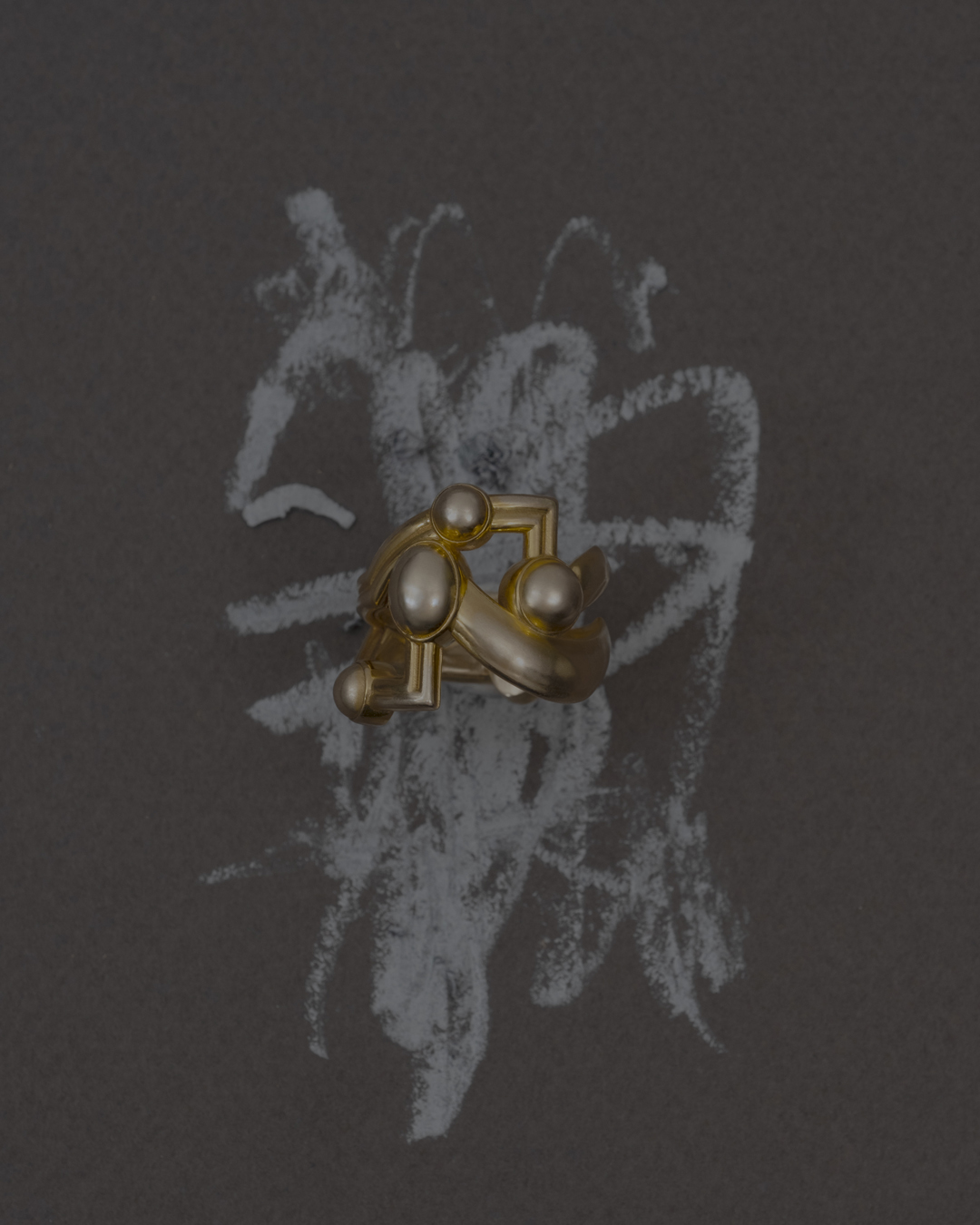 Abstract gold ring with sphere accents on a dark surface with chalky white scribbles in a minimalist still-life composition.
