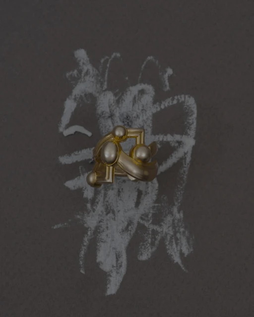 Abstract gold ring with sphere accents on a dark surface with chalky white scribbles in a minimalist still-life composition.