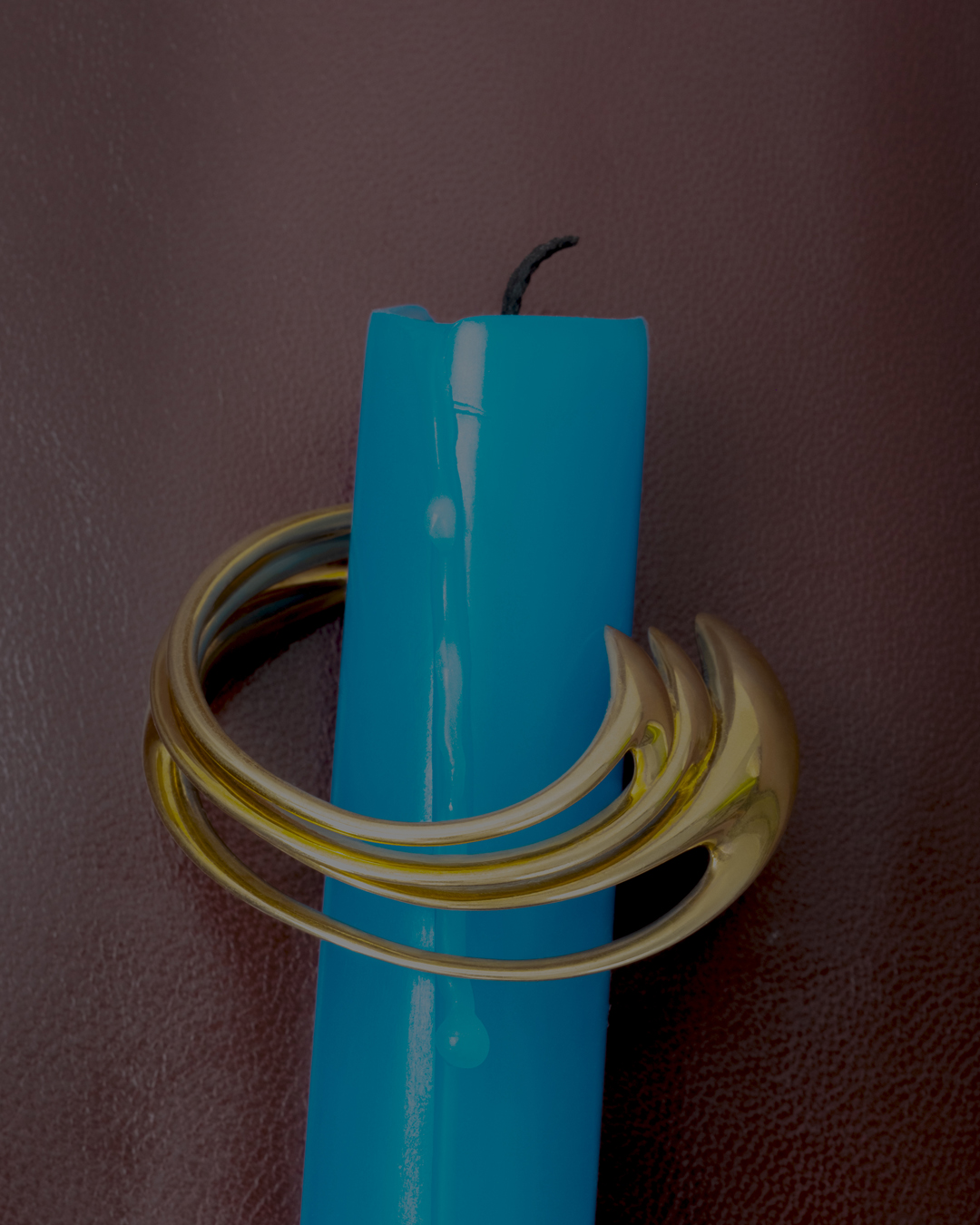 Close-up of a blue candle with a unique, curved gold metal candle holder, set against a dark brown backdrop.