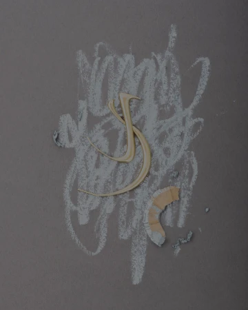 Stylized letter 'S' crafted from metal on a gray background with chalk scribble marks and broken pencil shaving, artful composition.