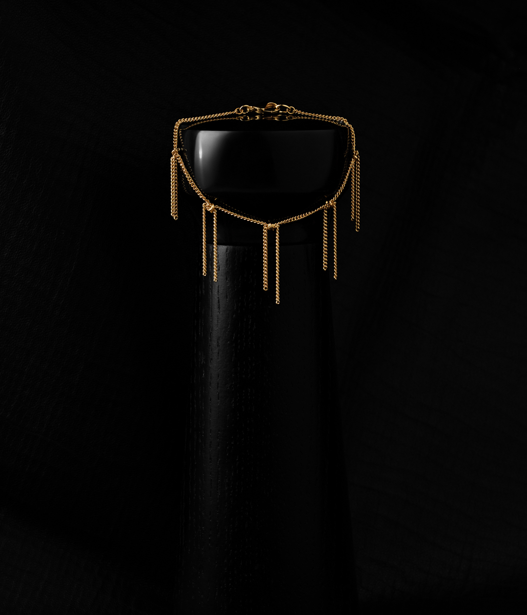Close-up of a gold fringe chain necklace draped over a black pillar against a dark background. Fashionable jewellery still life.