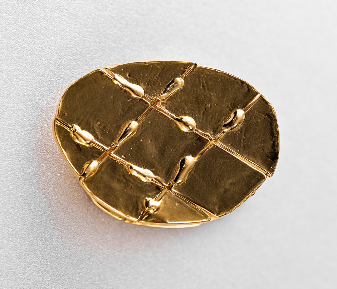 Elevated shot of a gold-toned decorative object with a grid pattern on a textured white surface, emphasizing metallic sheen and design.