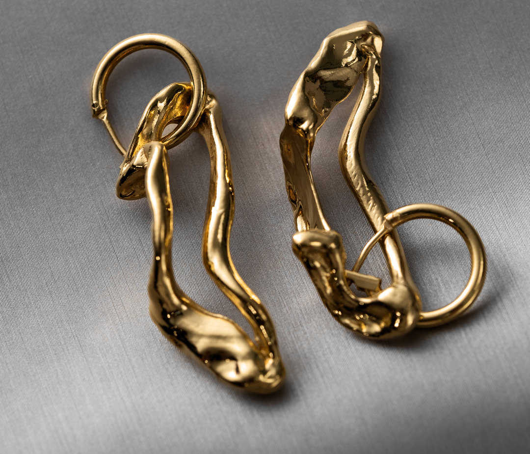 Close-up studio shot of two gold metal earrings with organic, flowing shapes on a light gray fabric background.