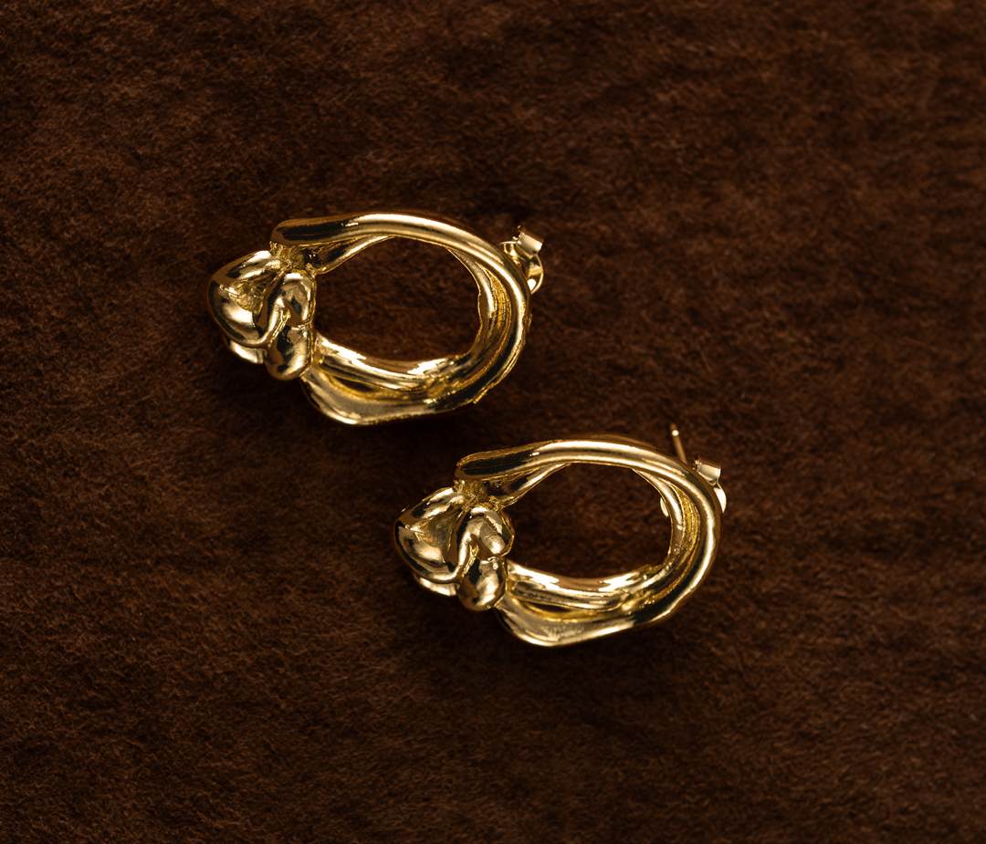 Close-up of two golden earrings on a brown textured surface, showcasing the jewelry's design and material in a simple composition.
