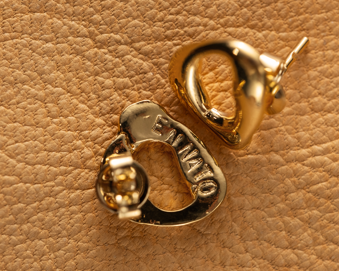 Close-up studio shot of two gold-toned metal earrings on a beige textured background, showcasing their heart-shaped loop design.