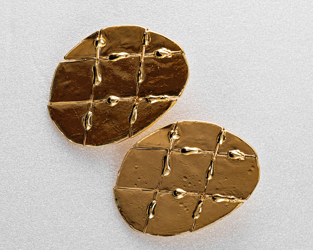 Overhead studio shot of two gold earrings with textured, geometric design on a white surface, showing detail and craftsmanship.