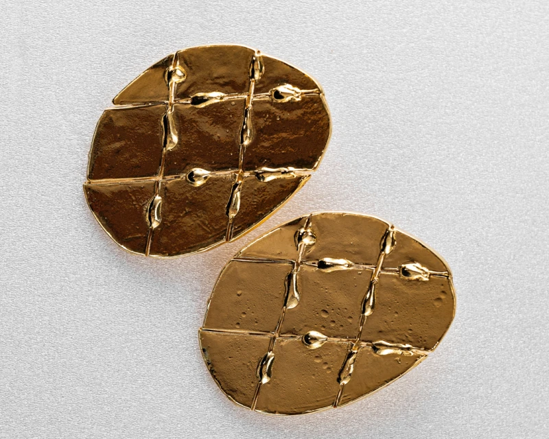 Overhead studio shot of two gold earrings with textured, geometric design on a white surface, showing detail and craftsmanship.
