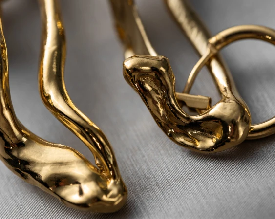 Close-up of sculpted organic gold jewellery with a molten finish resting on a textured grey fabric surface.