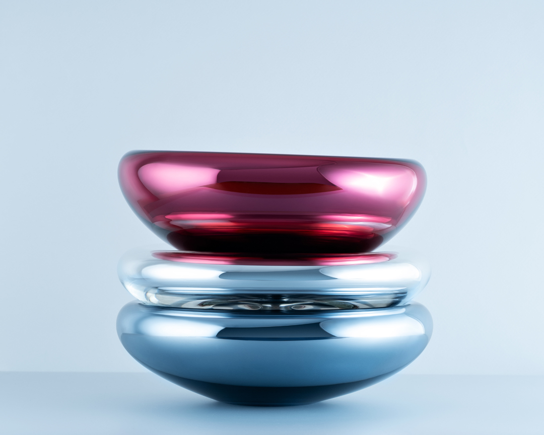 Still life of stacked glass bowls in red, silver, and blue on a soft blue background with studio lighting showcasing their curves and reflections.
