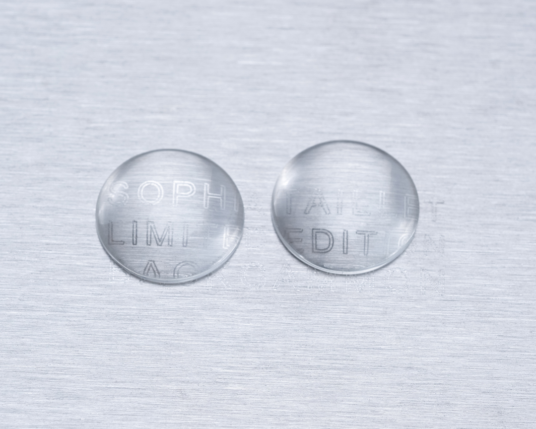 Two clear, round objects with etched text on a brushed metal surface, creating a minimalist and modern abstract composition.