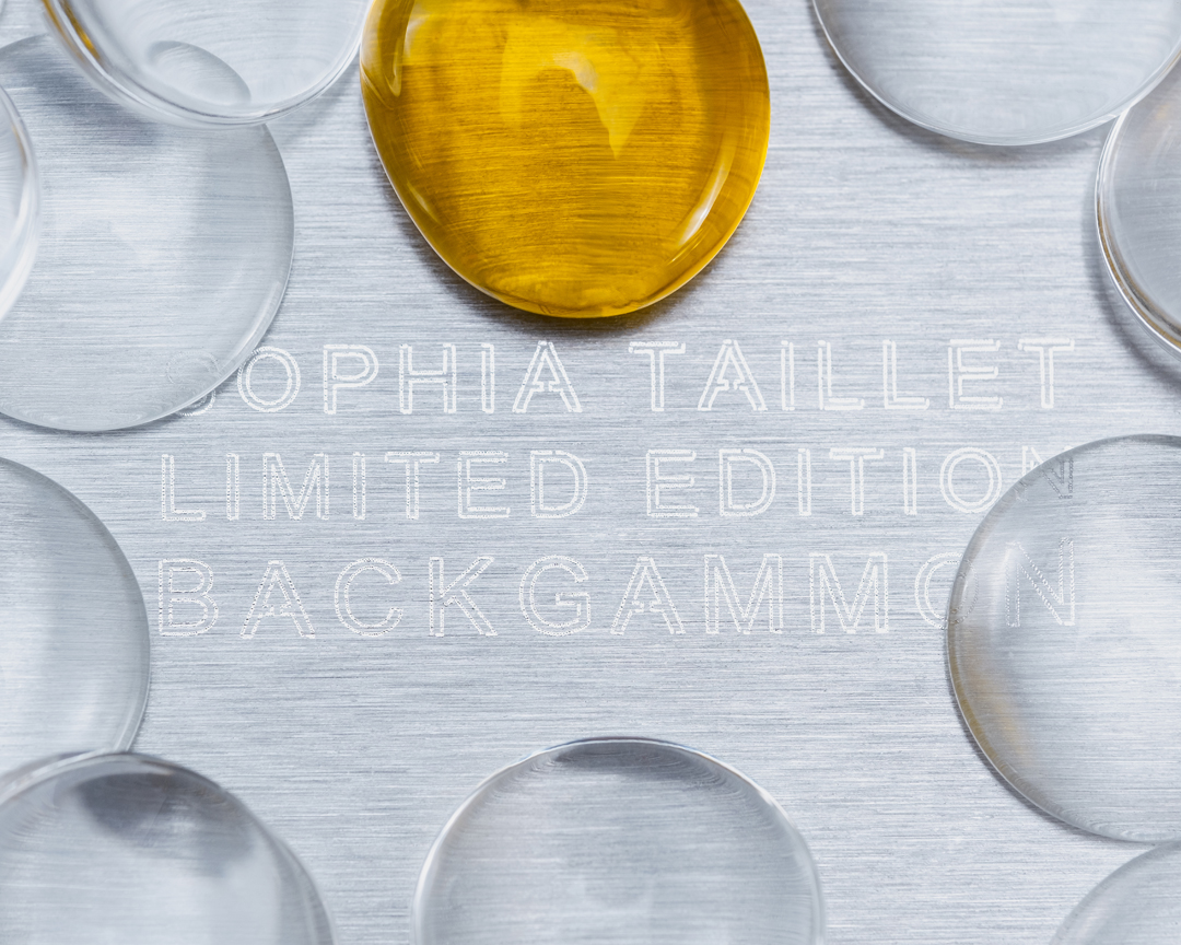Overhead shot of a backgammon game board with transparent and gold game pieces arranged on a silver surface etched with text.