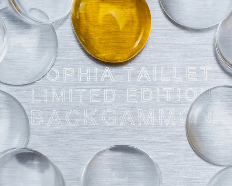 Overhead shot of a backgammon game board with transparent and gold game pieces arranged on a silver surface etched with text.