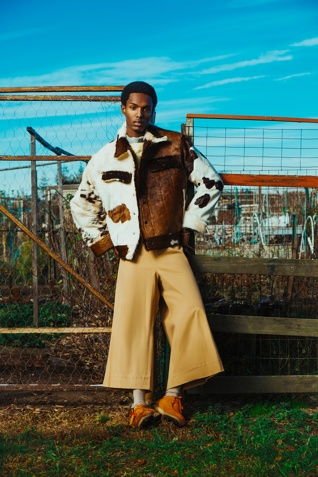 A man wearing a cow-print shearling jacket and wide trousers standing before a wire fence under a blue sky.