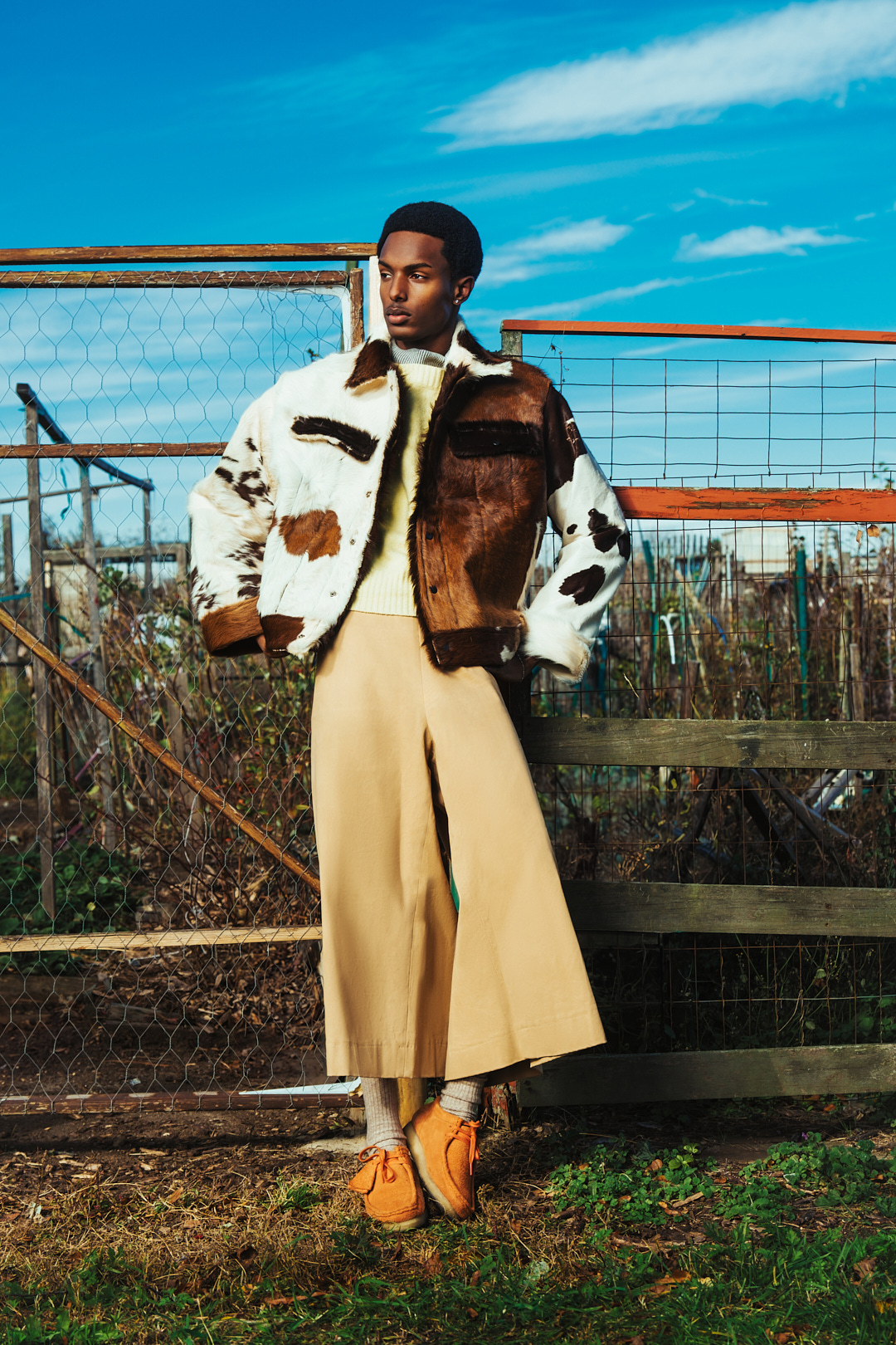 A dark-skinned male model poses in sunlight against a rusty fence, wearing a fur jacket, tan culottes, and orange moccasins in a grassy field.
