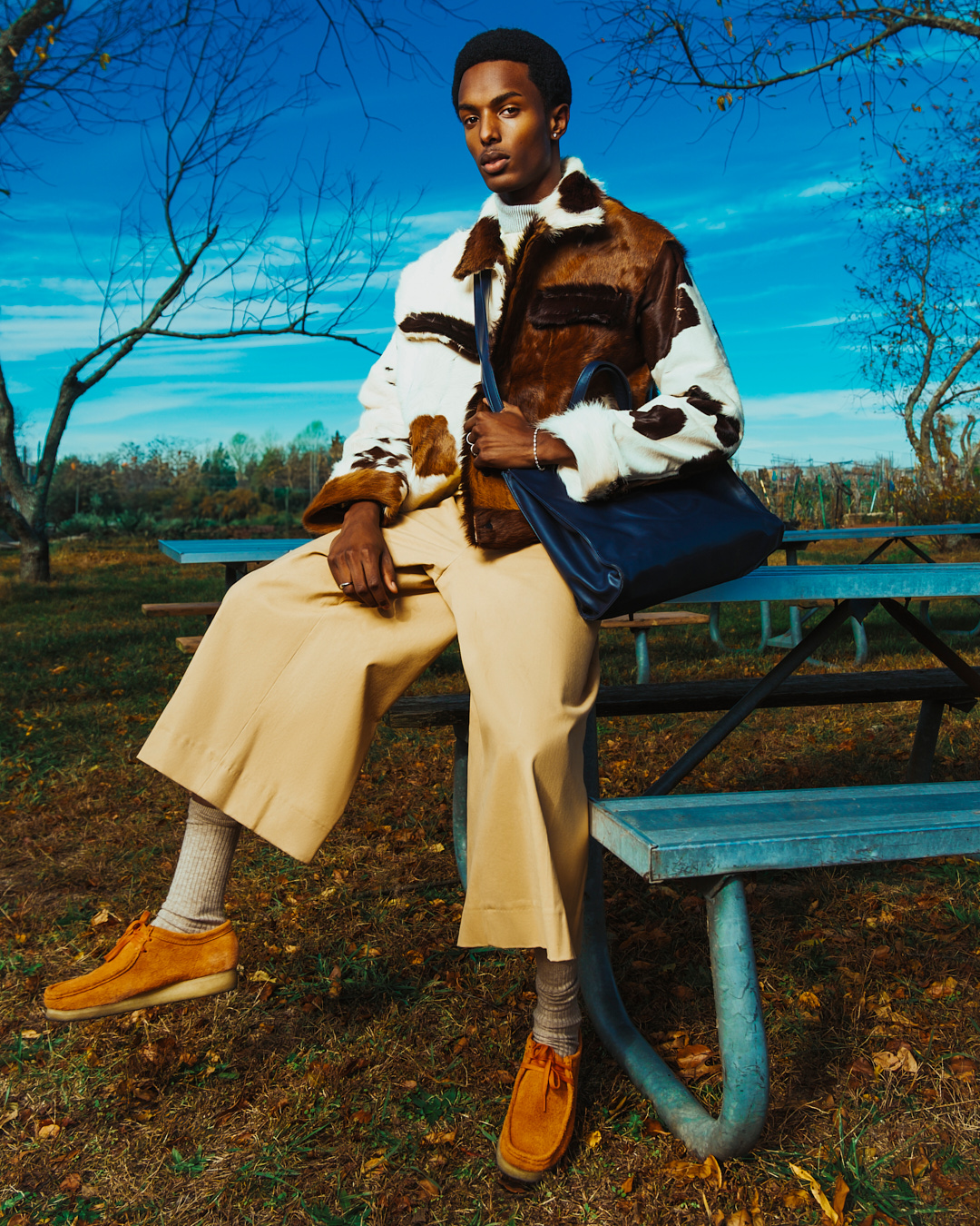 Man in fur jacket and tan pants sits on a blue picnic table outside, accessorized with a shoulder bag and suede shoes against a blue sky.