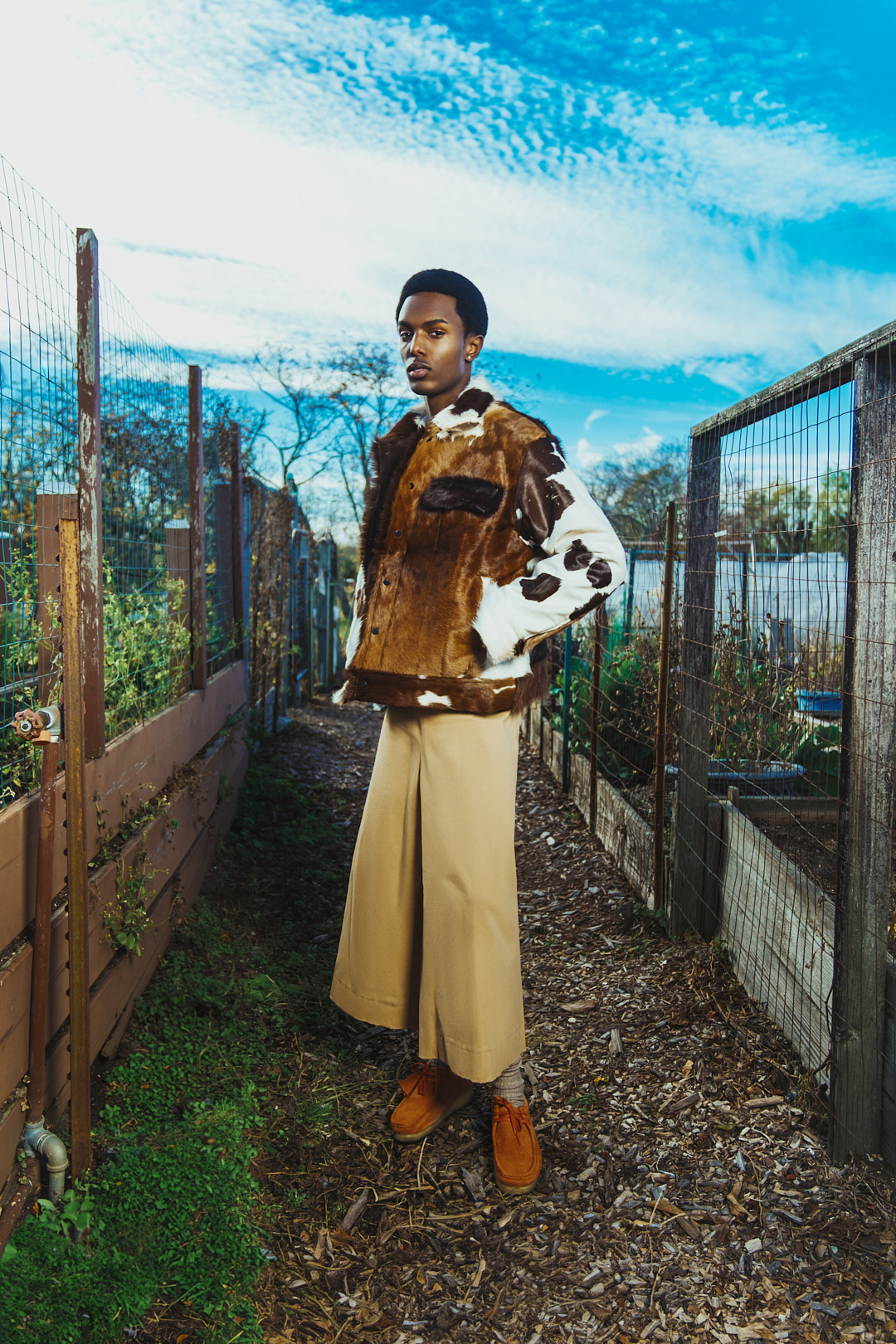 Full shot of a Black male model standing on a garden path wearing a brown and white cowhide jacket, tan pants, and brown shoes.
