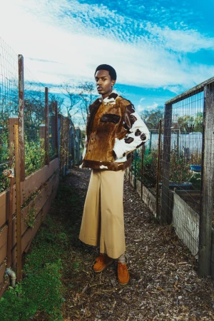 Full shot of a Black male model standing on a garden path wearing a brown and white cowhide jacket, tan pants, and brown shoes.