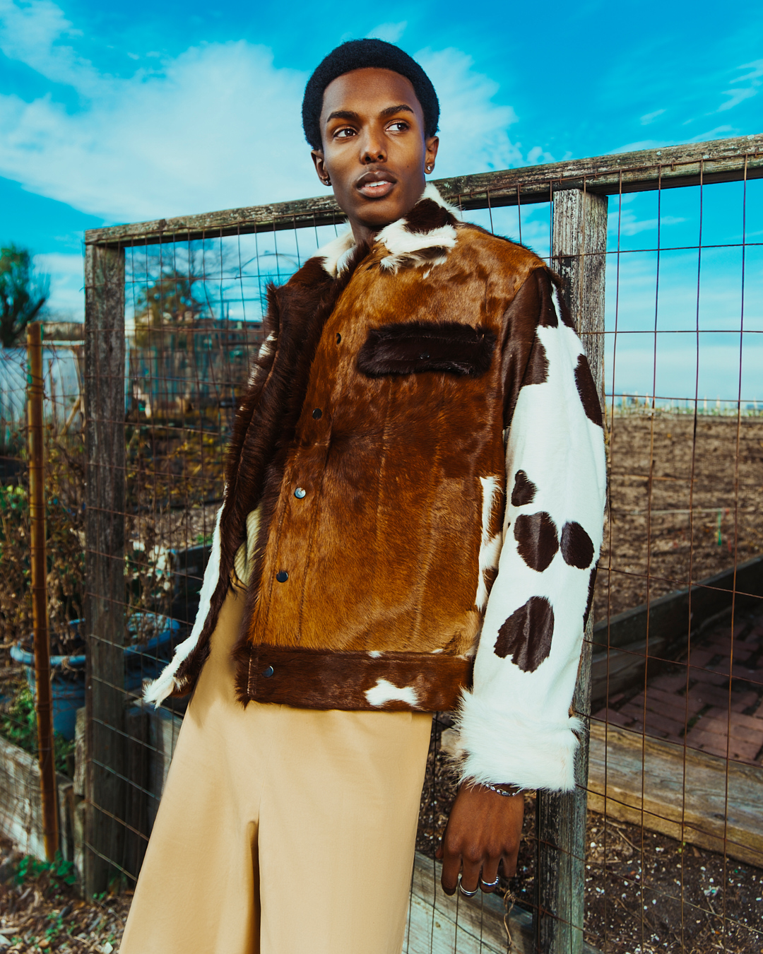 Portrait of a young Black man wearing a fur-trimmed brown jacket in an outdoor setting with a wooden fence and blue sky.