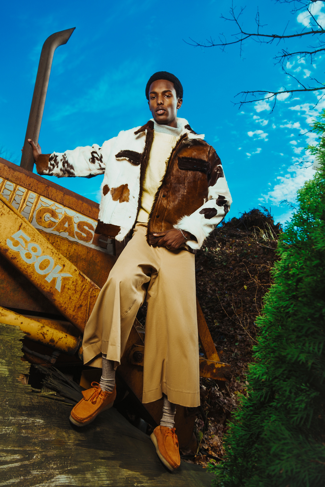 Man in patterned jacket poses next to construction equipment under blue sky. Fashion editorial shot featuring outdoor setting.