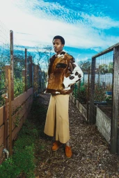 A man standing on a garden path wearing a brown and white cowhide jacket and wide-leg beige trousers under a blue sky.