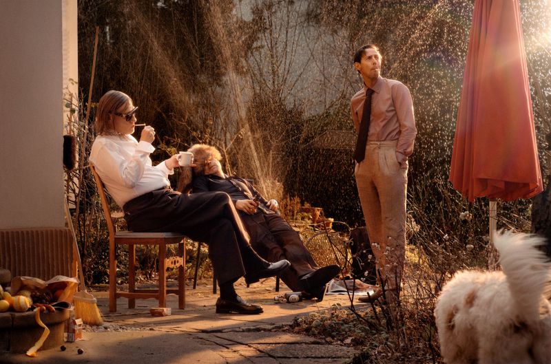 Three men on a patio, one standing wearing a tie, one seated smoking, and one reclining with a drink, with a dog in the foreground.
