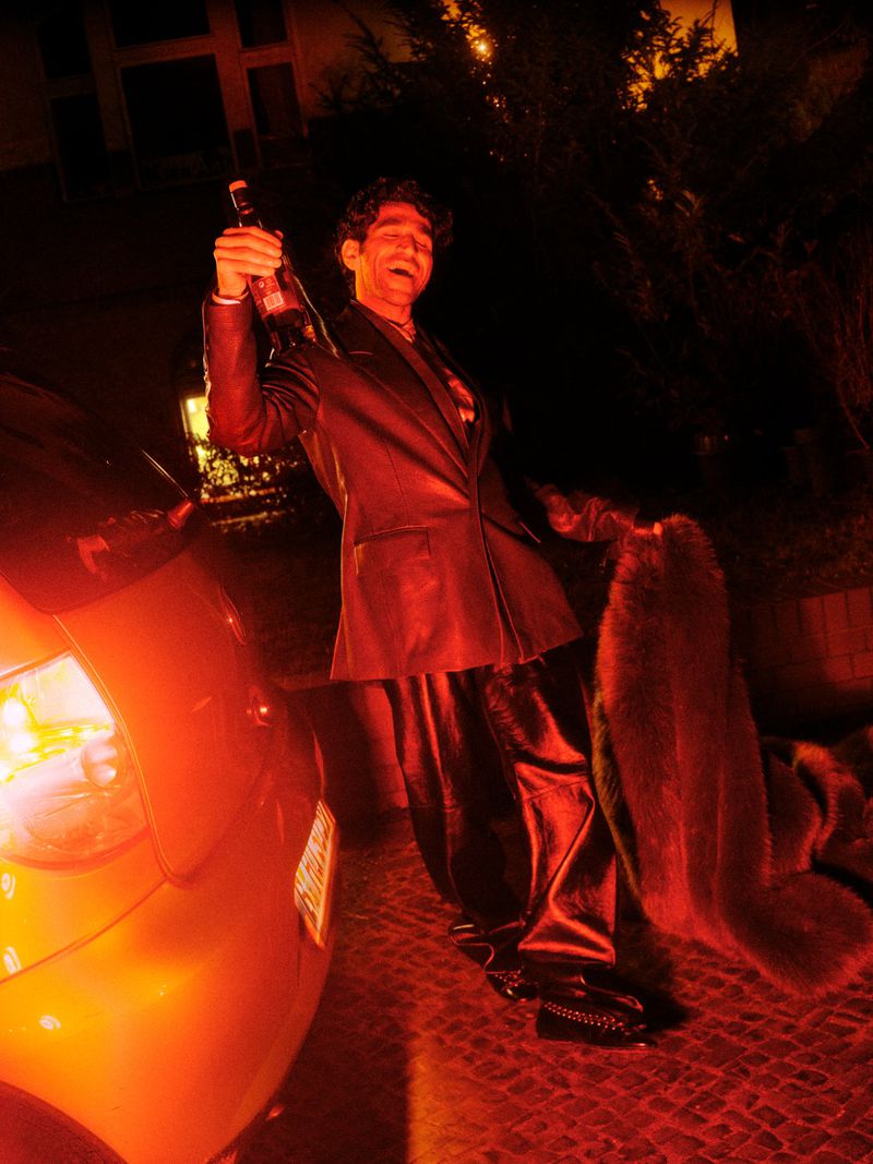 A man in a black suit and fur coat holds a champagne bottle next to a parked car under a red light at nighttime.