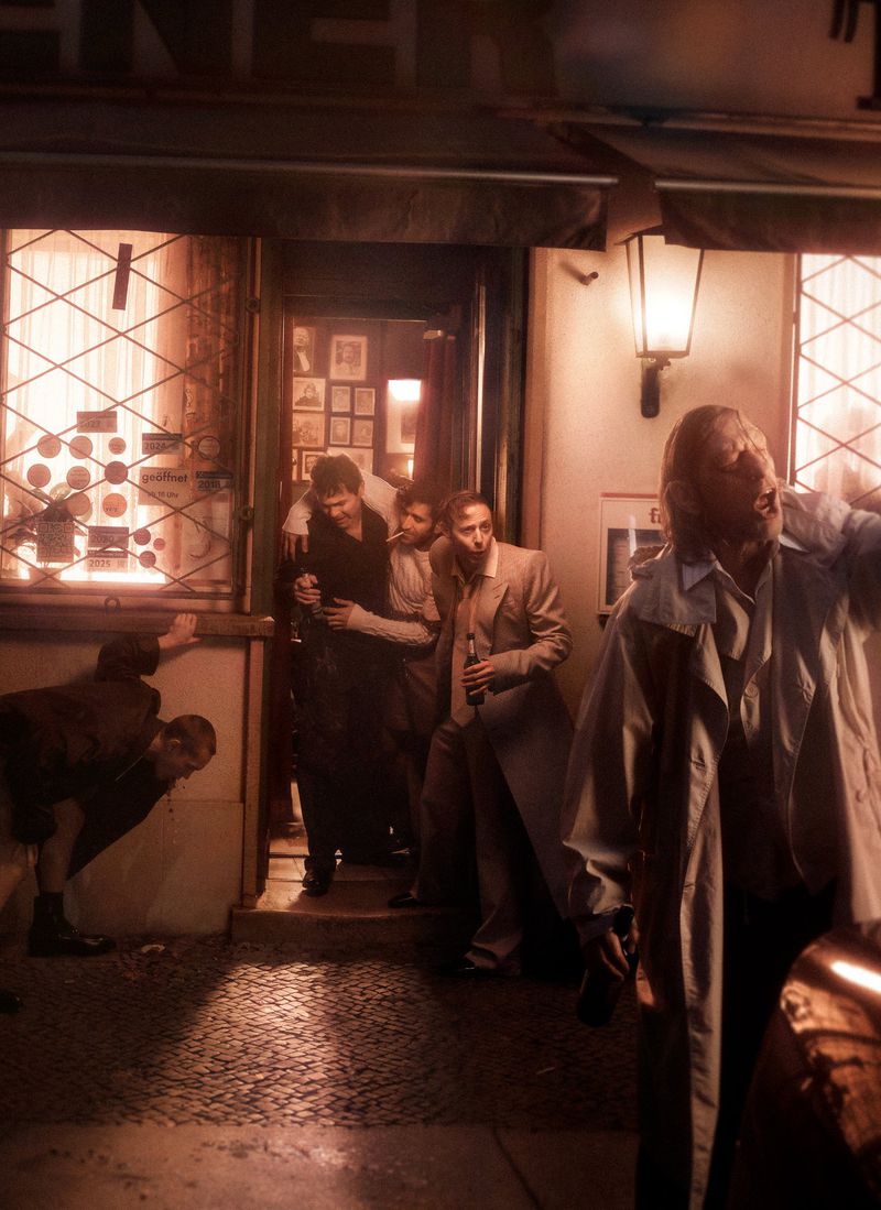 Outdoor night shot of a group of men standing outside a building entrance, some holding bottles, in an urban environment.