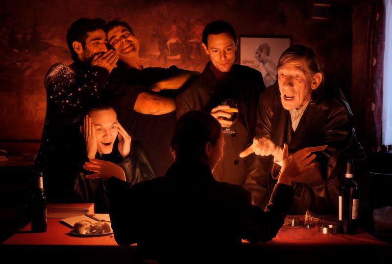 Group of people gathered at a table with wine and snacks, engaged in conversation and laughter in a dimly lit indoor setting.