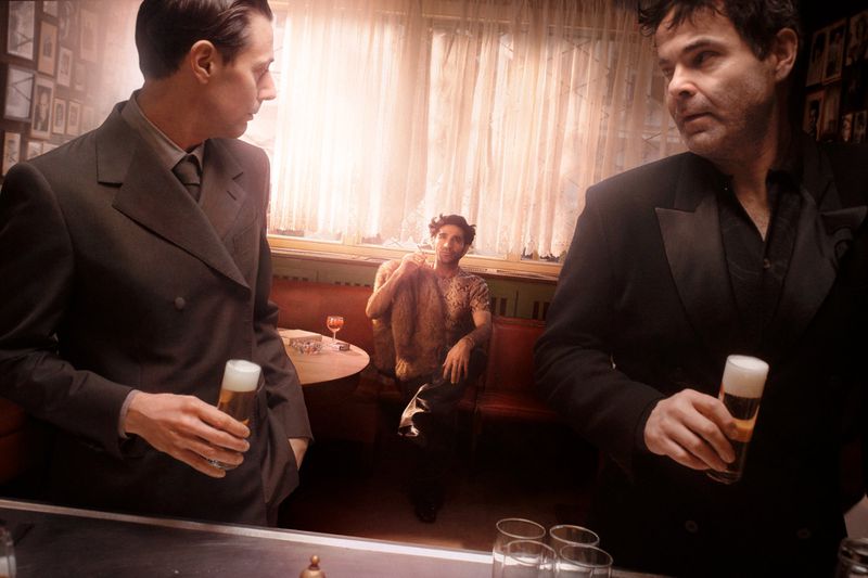 Three men in suits are in a dimly lit bar setting, one seated in the background, holding glasses with beer, creating a vintage atmosphere.