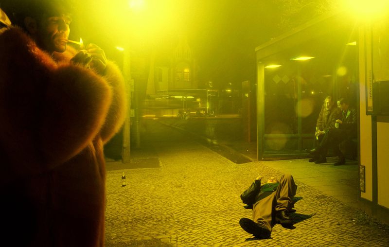 A man in a fur coat is lighting a cigarette in an urban nighttime scene. Another man lies on the ground. Bus stop with people in background.