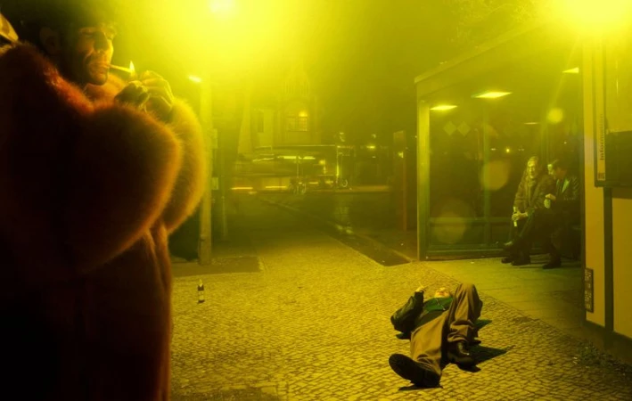 A man in a fur coat is lighting a cigarette in an urban nighttime scene. Another man lies on the ground. Bus stop with people in background.