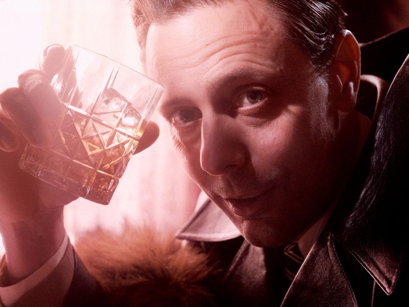 Adult man in a dark jacket raises a faceted glass of amber liquor with ice in an indoor setting with diffused lighting.
