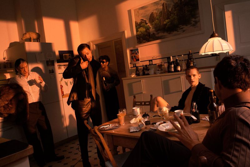 Indoor gathering featuring six adults in casual clothing around a wooden table with bottles, cups, and plates in a warm-toned room.