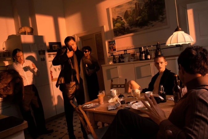 Indoor gathering featuring six adults in casual clothing around a wooden table with bottles, cups, and plates in a warm-toned room.