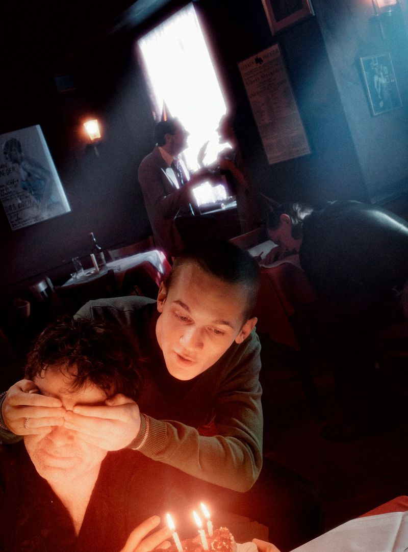 Man covering another man's eyes next to a small cake with lit candles, other people in the background in a dimly lit interior.